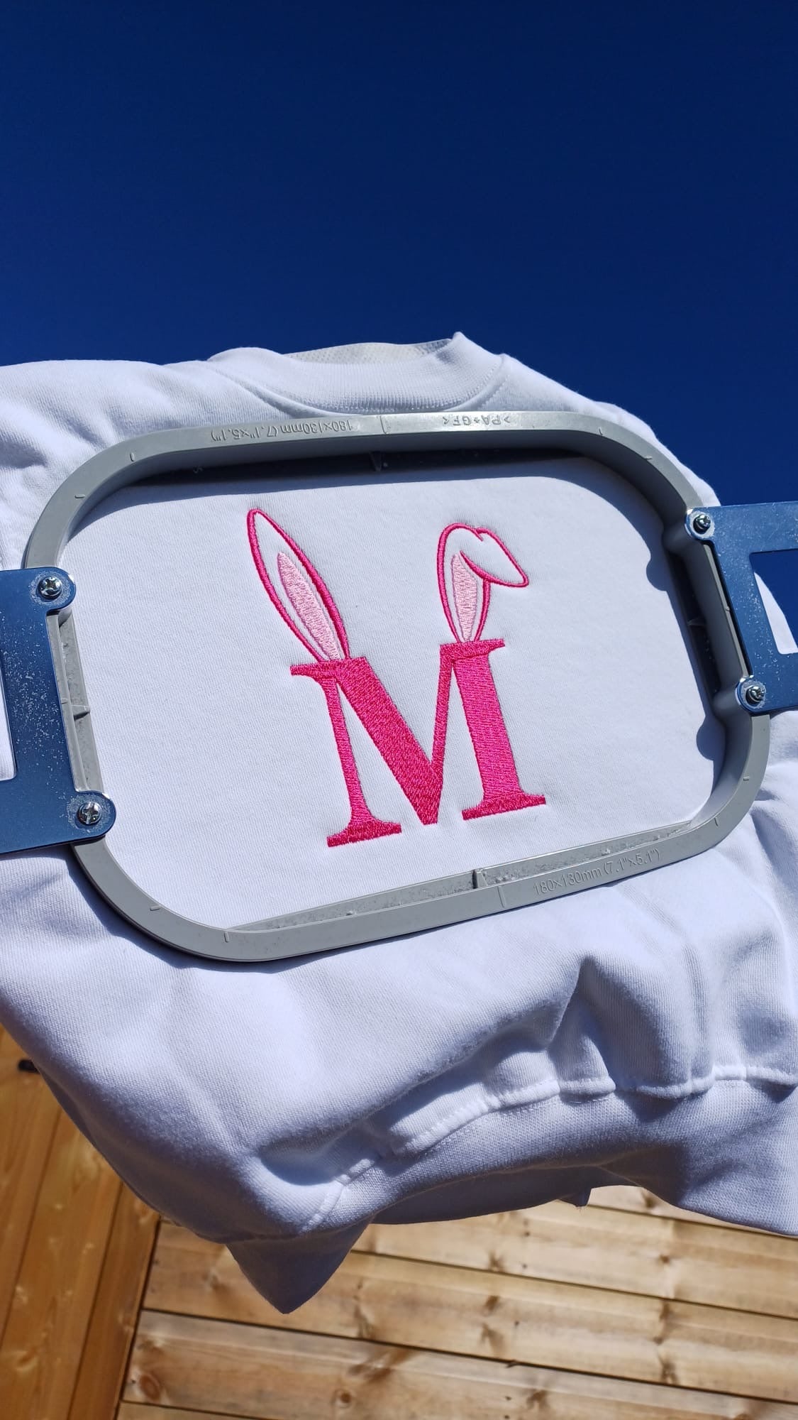 Embroidery hoop with pink letter 'M' design on a white fabric against a blue sky.