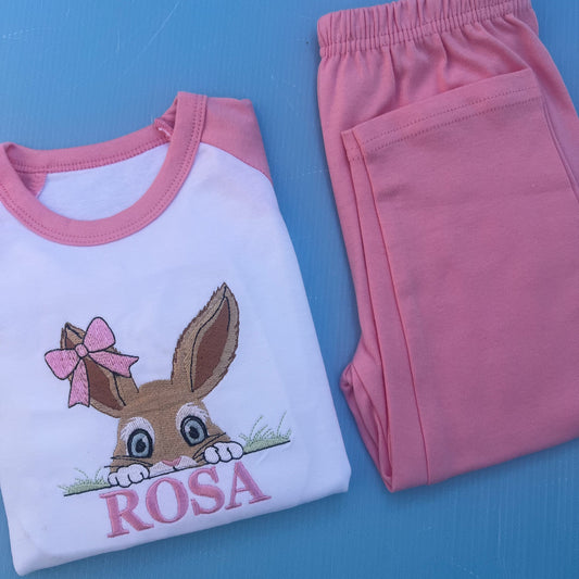 Pink pyjamas with a bun peaking over some grass with a bow on her ear. Finished off with the name Rosa.
