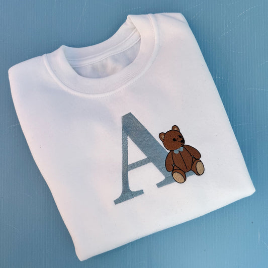 White t-shirt with a brown teddy bear and letter 'A' on a blue background