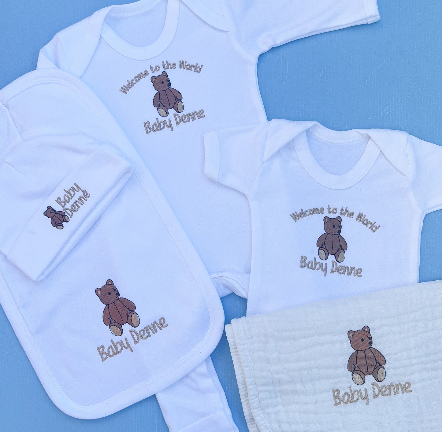 Baby clothing set with embroidered teddy bear and text on a blue background