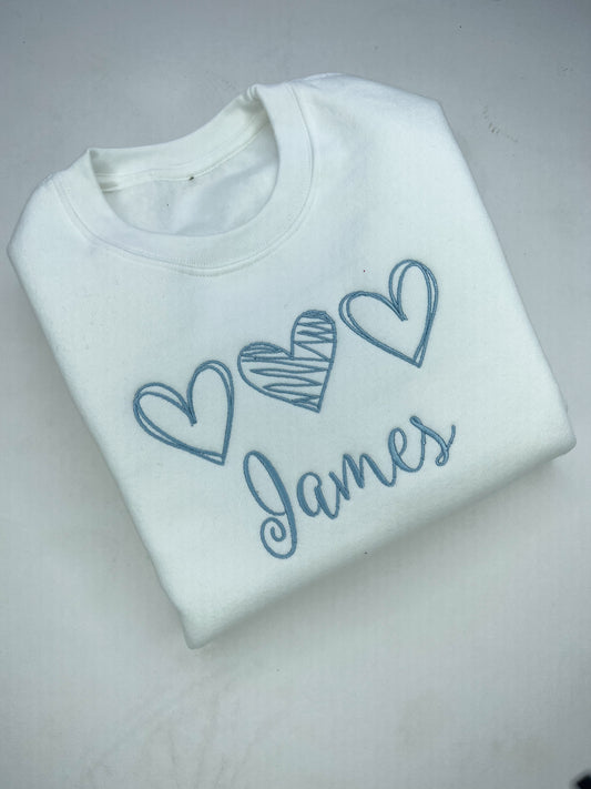 White sweatshirt with blue hearts and 'James' text on a light gray background