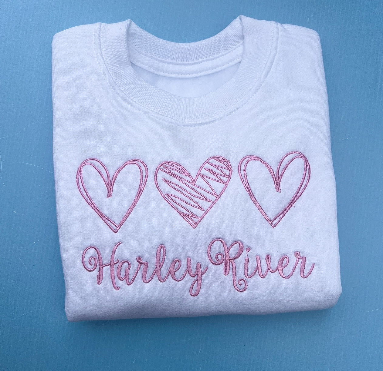 Folded white sweatshirt with pink embroidered hearts and 'Harley River' text on a blue background