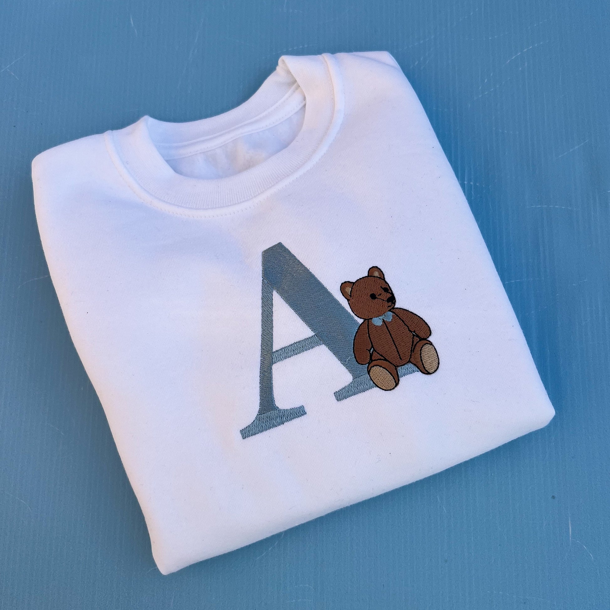 White sweatshirt with a teddy bear and letter 'A' on a blue background