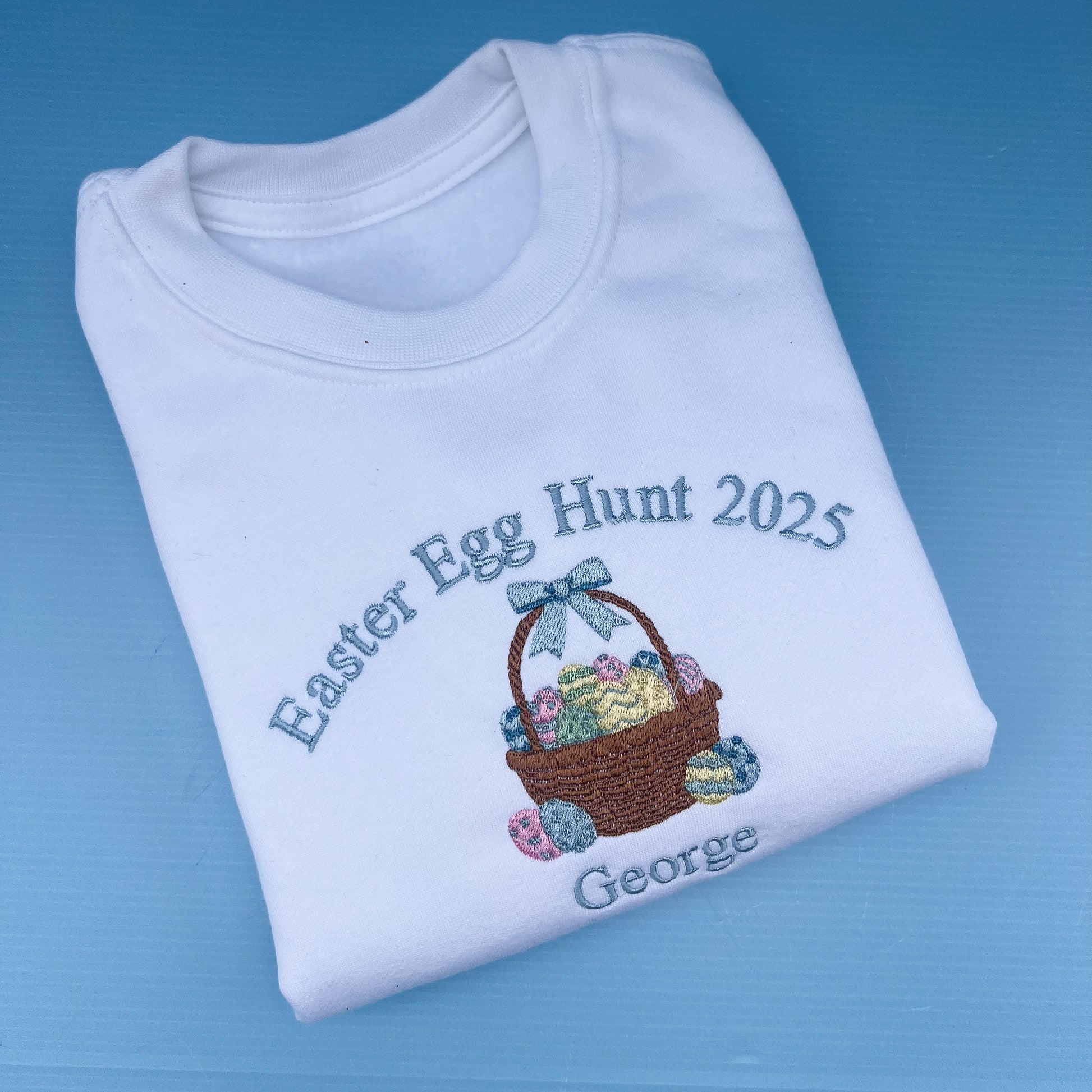 Folded white sweatshirt with Easter Egg Hunt 2025 design on a blue background