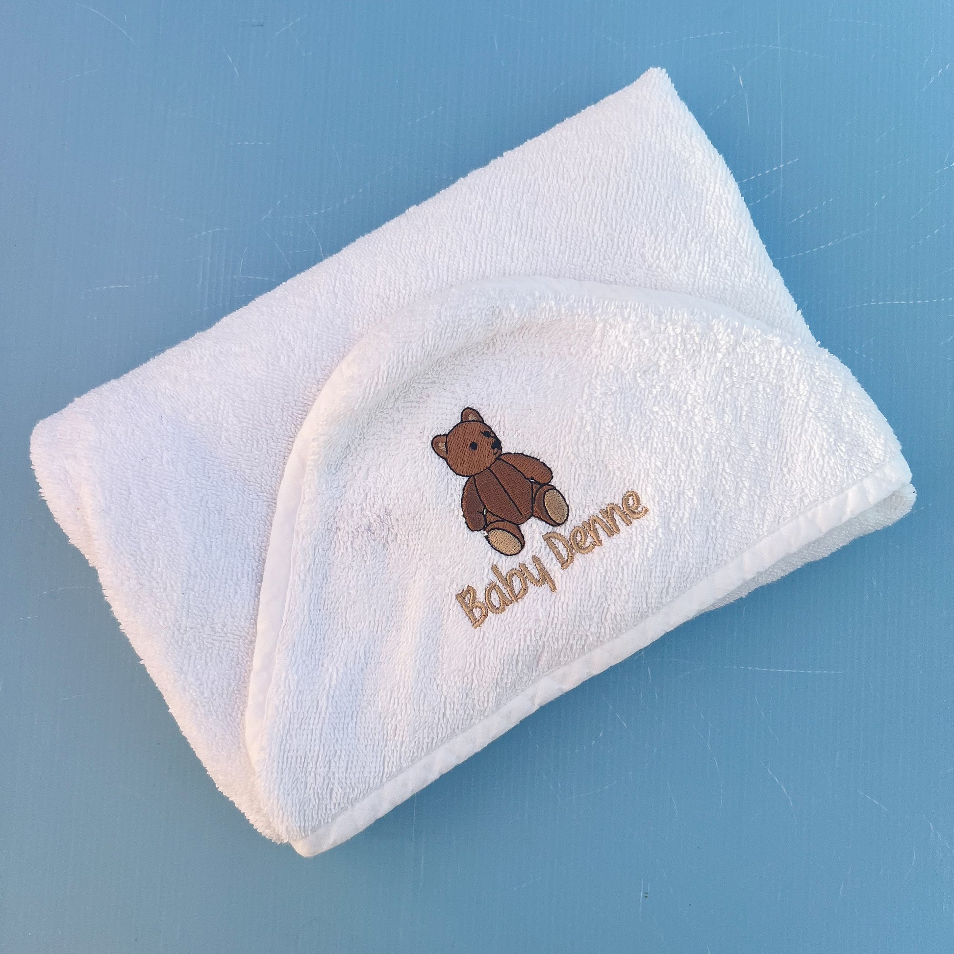 White baby towel with brown teddy bear design and 'Baby Denne' text on a blue background