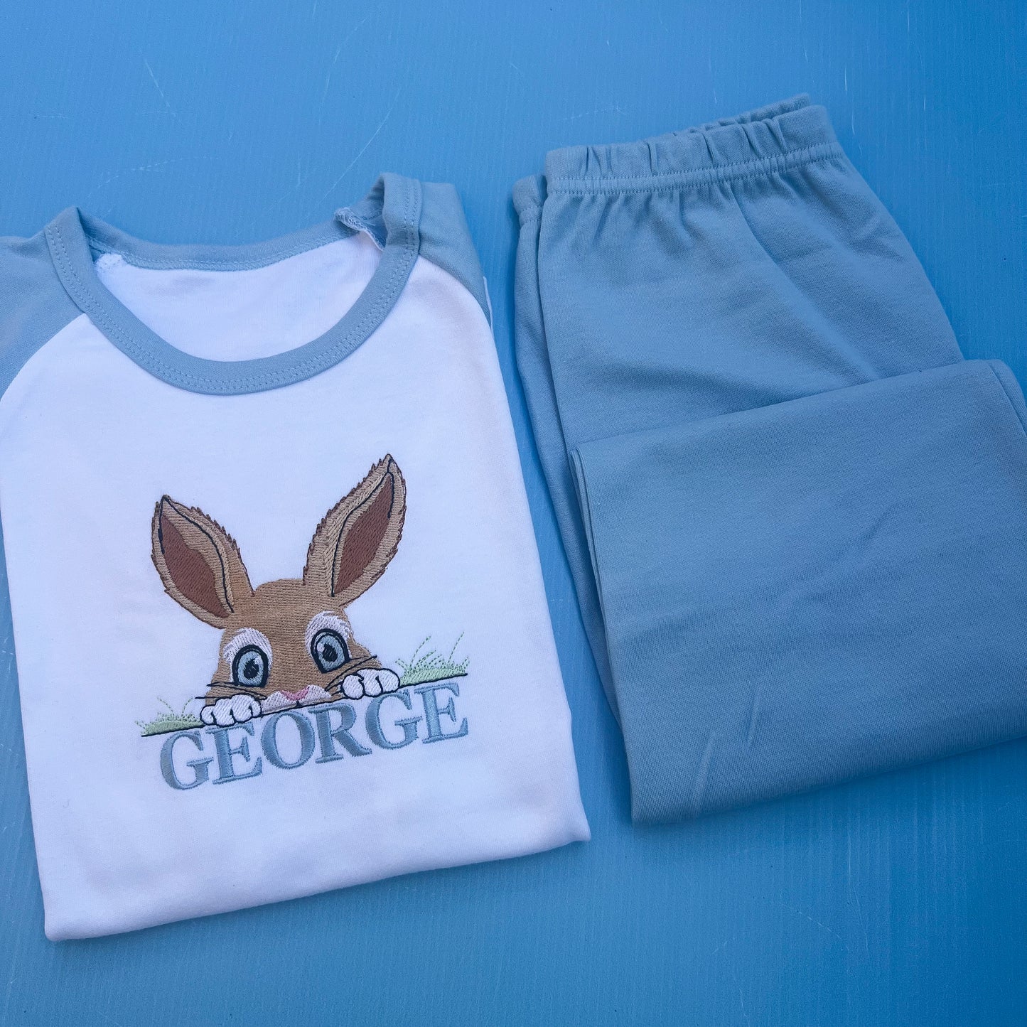 Blue pyjamas with a peaking bunny posting over grass embroidered on with the name George 