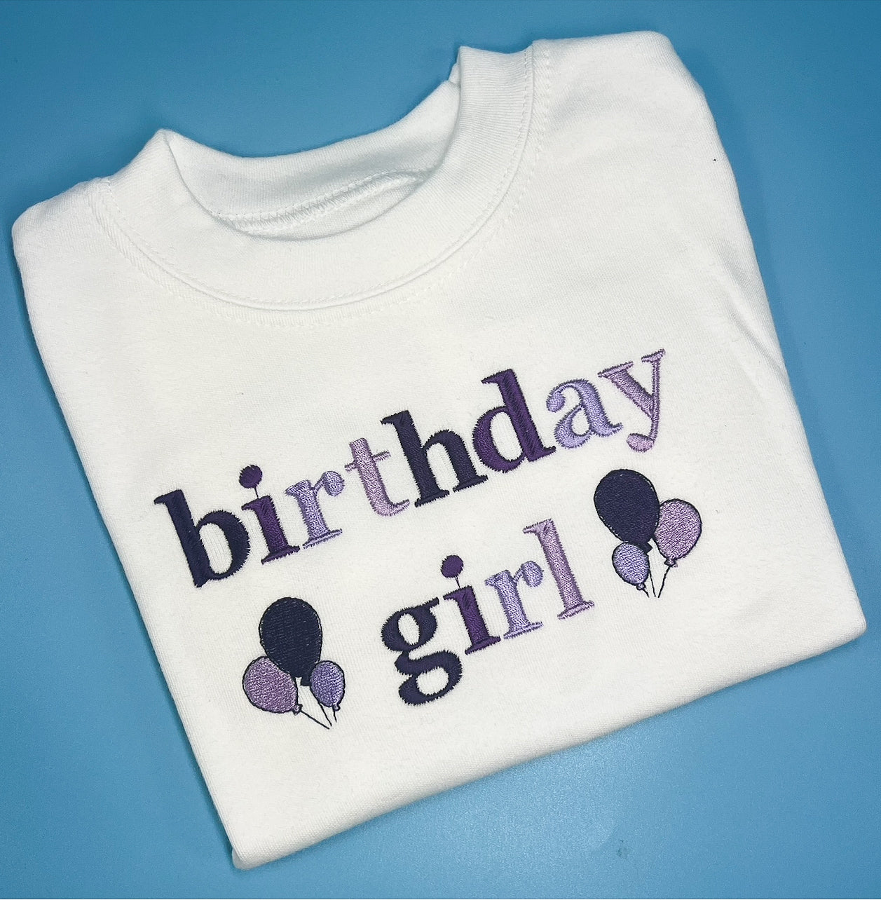 Folded white sweatshirt with 'birthday girl' text and balloons on a blue background