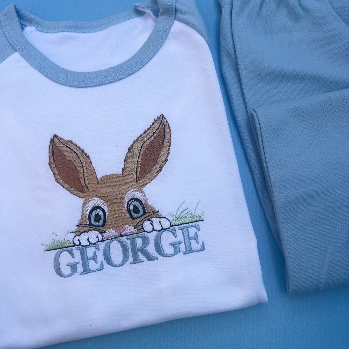 Blue pyjamas with a peaking bunny posting over grass embroidered on with the name George 