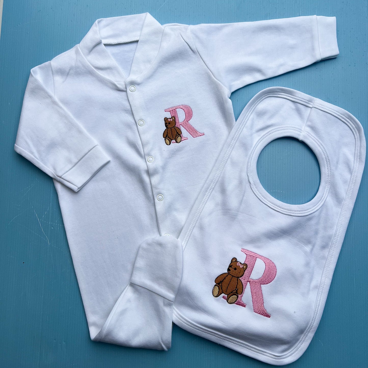 White baby outfit with embroidered teddy bear and 'R' on a blue background