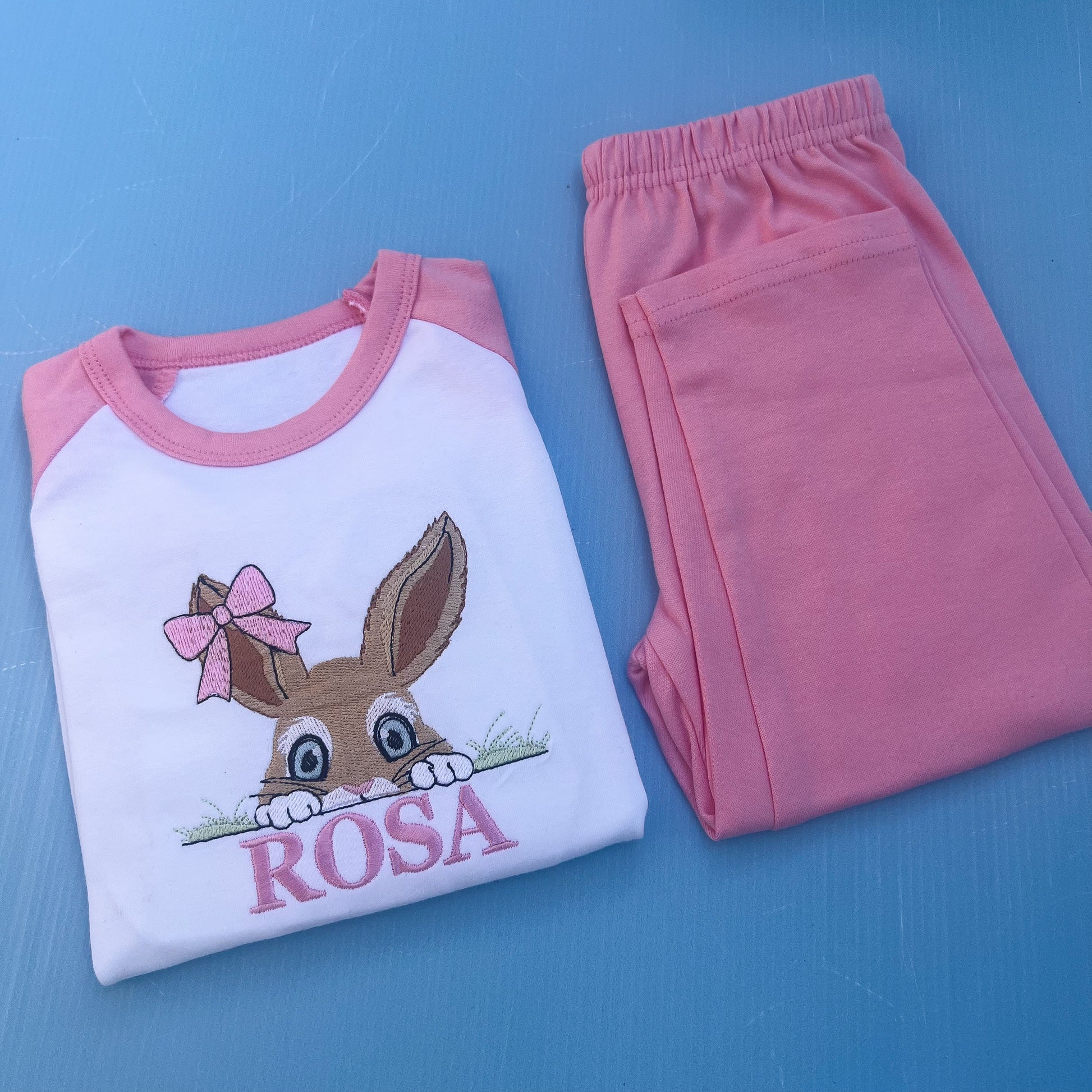 Pink pyjamas with a bun peaking over some grass with a bow on her ear. Finished off with the name Rosa. 