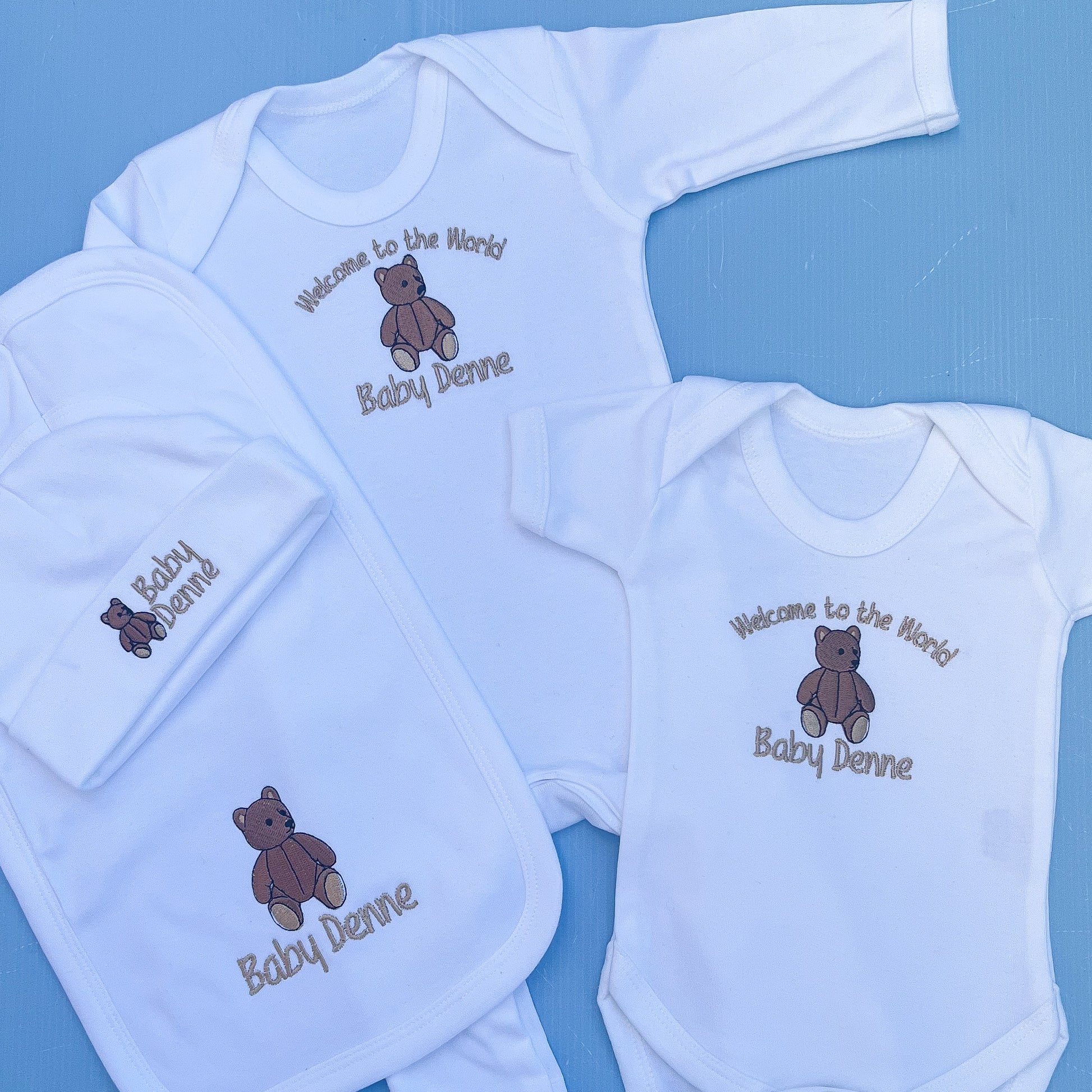 Baby clothing set with embroidered text and teddy bear design on a blue background