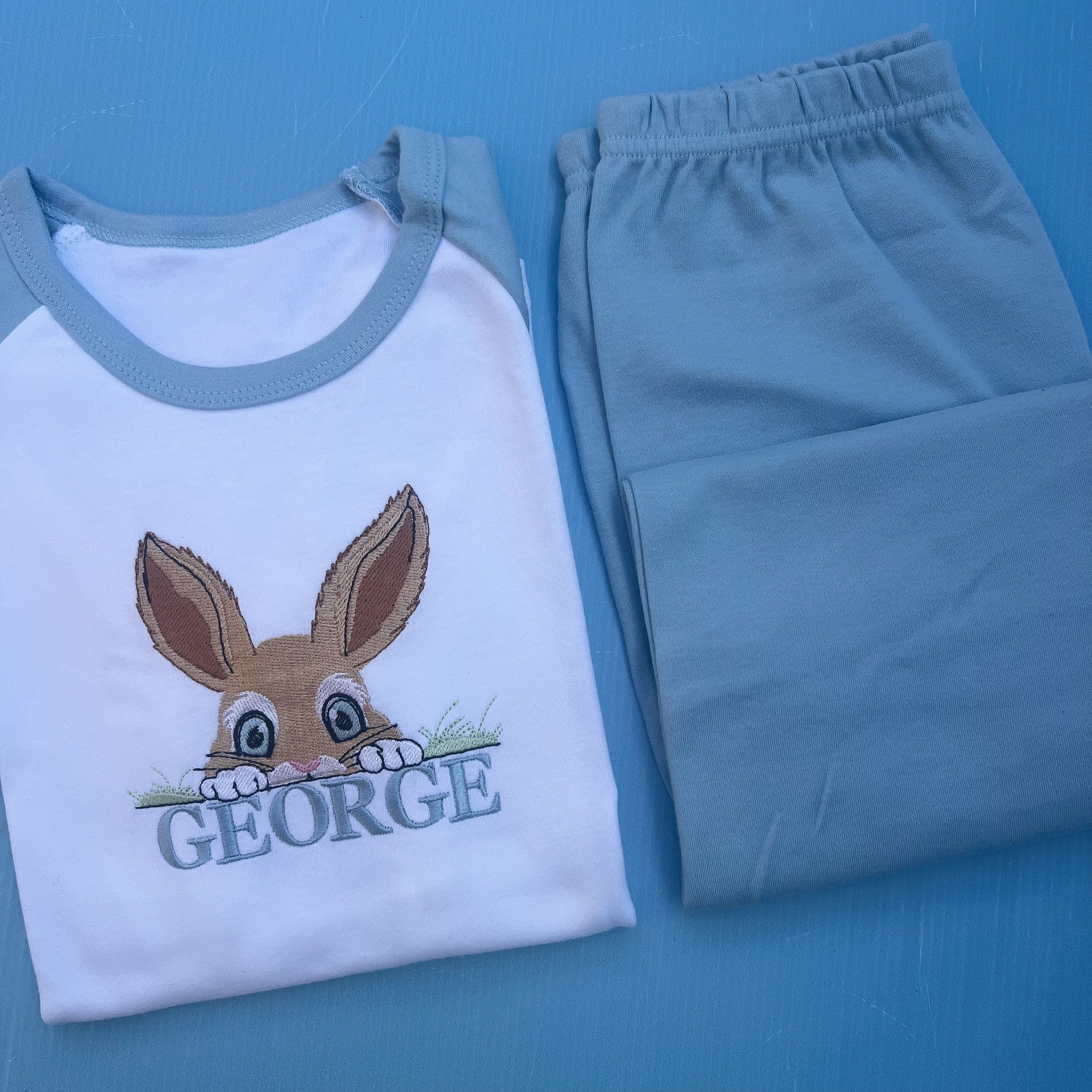 Blue pyjamas with a peaking bunny posting over grass embroidered on with the name George 