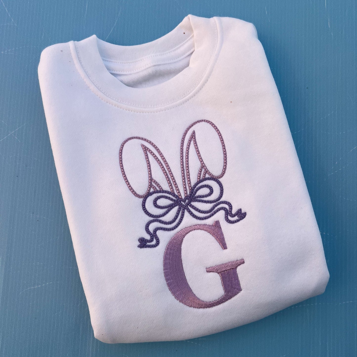 White jumper with embroidered bunny ears, a large bow and the letter G 
