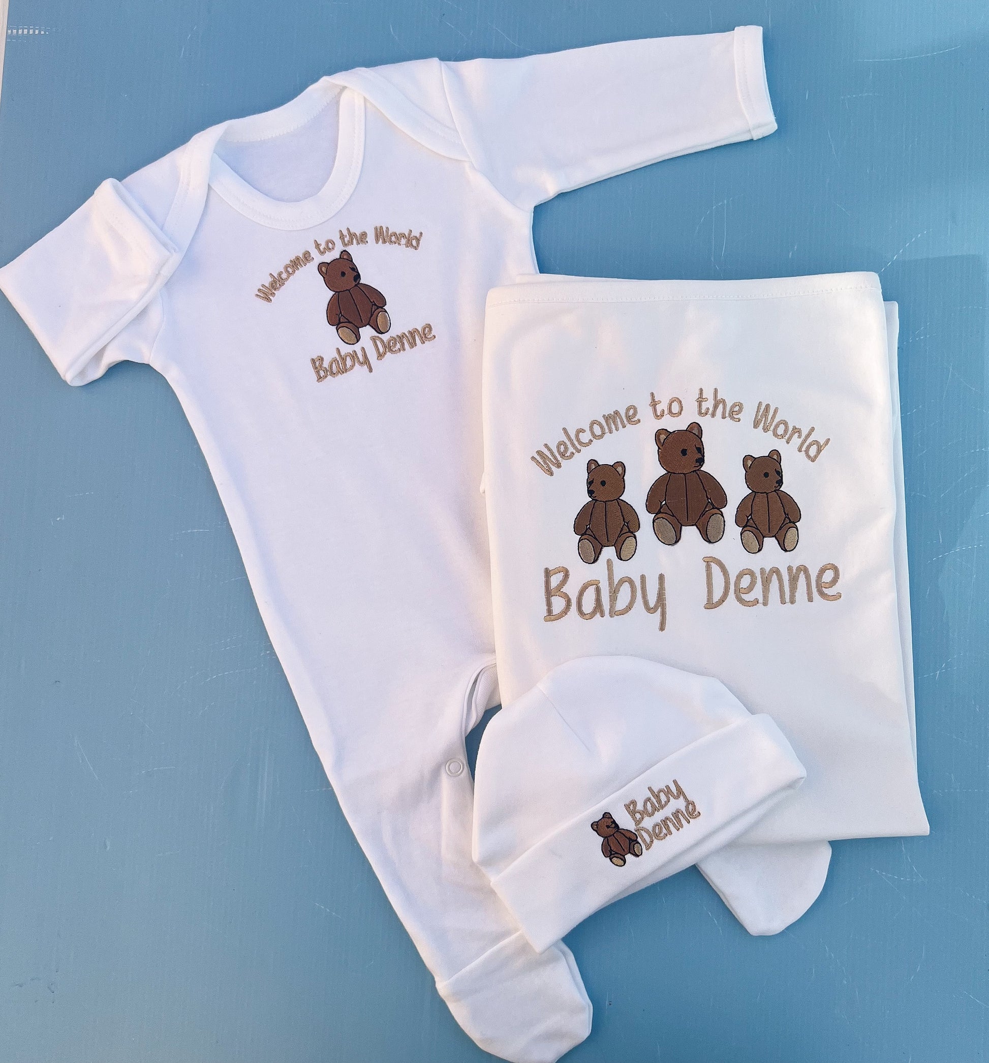 Baby clothing set with 'Welcome to the World Baby Denne' text and teddy bear graphics on a blue background.
