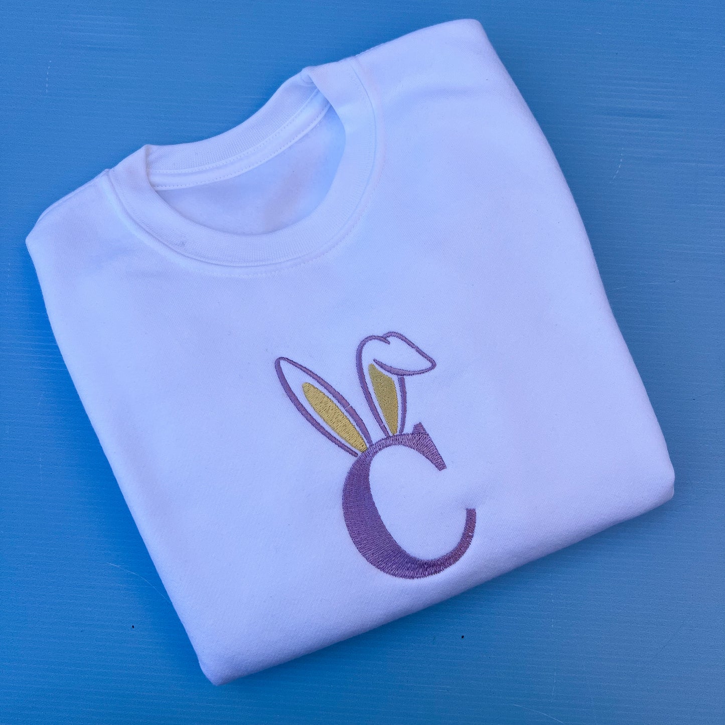Folded white sweatshirt with a purple letter 'C' and bunny ears on a blue background