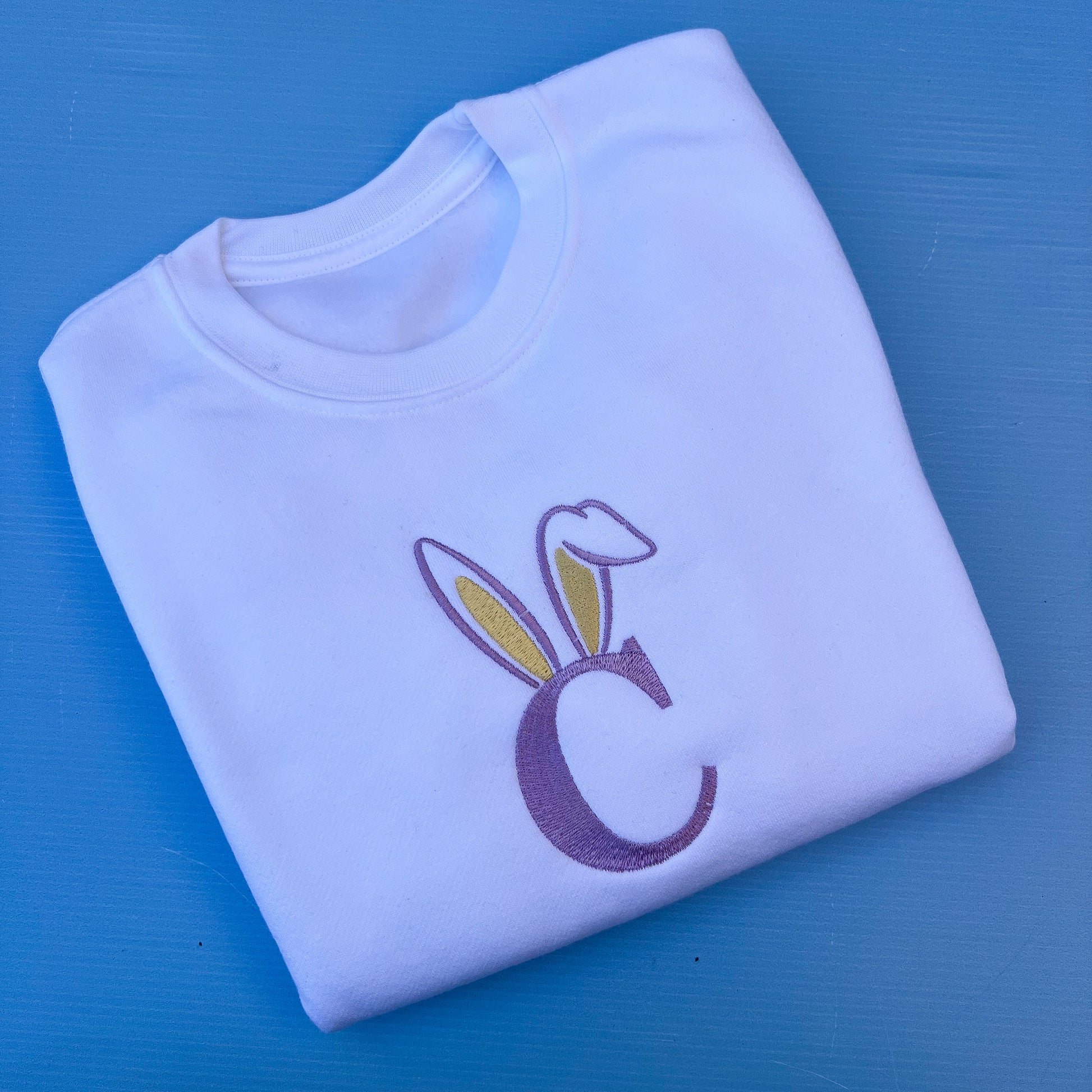 Folded white sweatshirt with a purple letter 'C' and bunny ears on a blue background