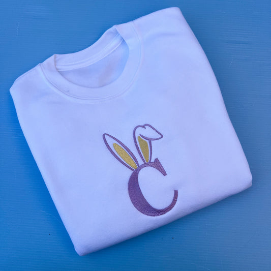 Folded white sweatshirt with a purple letter 'C' and bunny ears on a blue background