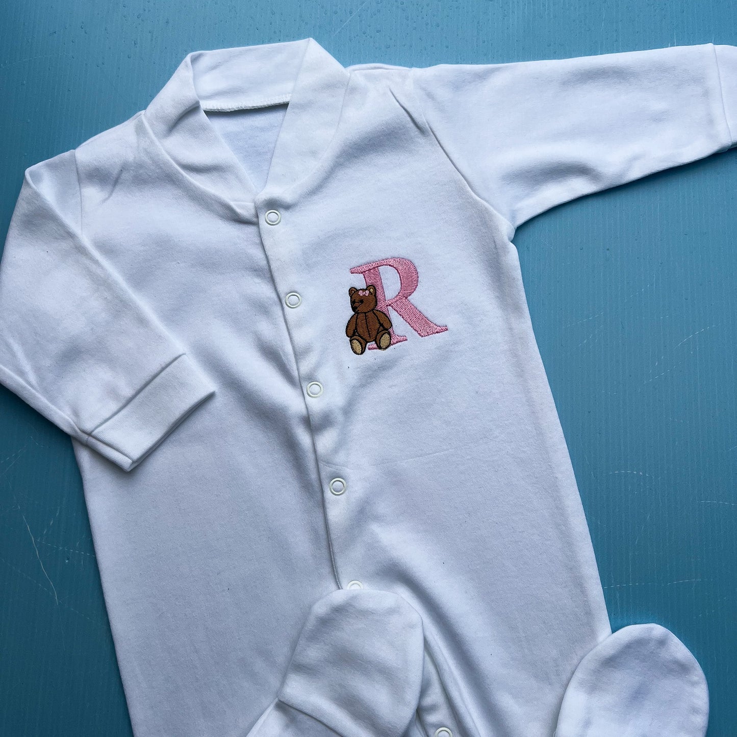 White baby onesie with a teddy bear and letter 'R' on a blue background