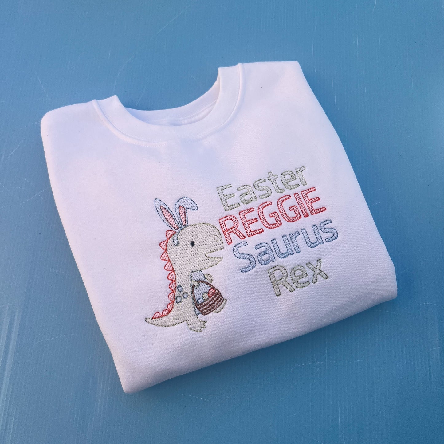 White sweatshirt with a dinosaur with bunny ears and a Easter egg basket with the words Easter Reggie Saurus Rex 