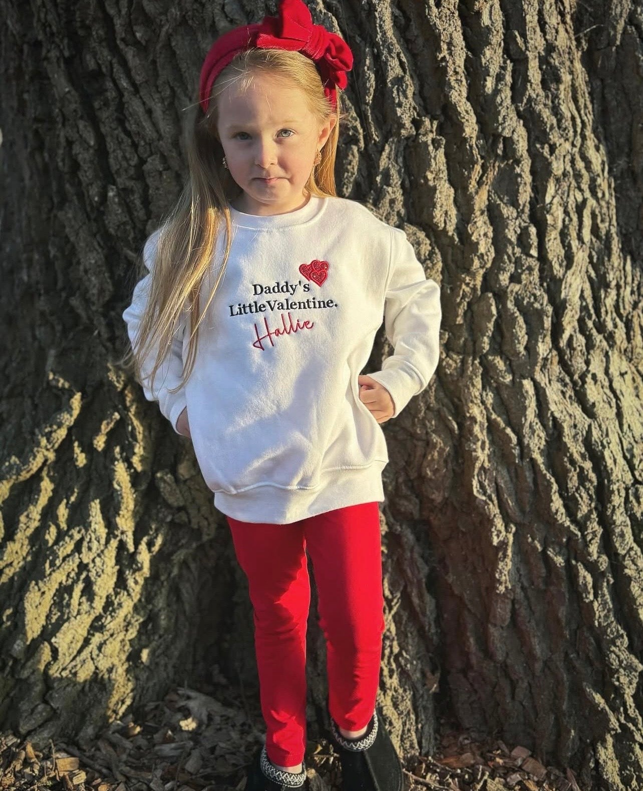 Young girl wearing a white sweatshirt with text and red pants standing against a tree. Daddy’s little Valentine sweatshirt 