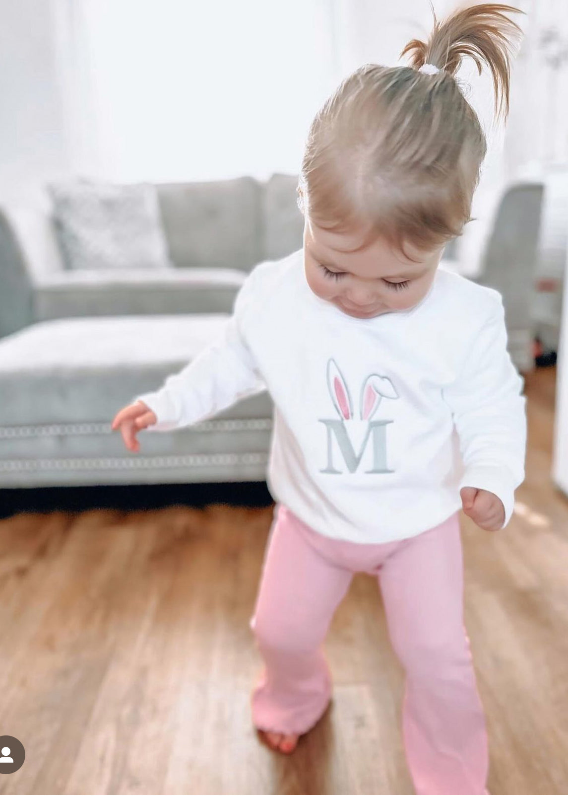 Child wearing a white shirt with a bunny design and pink pants in a living room.