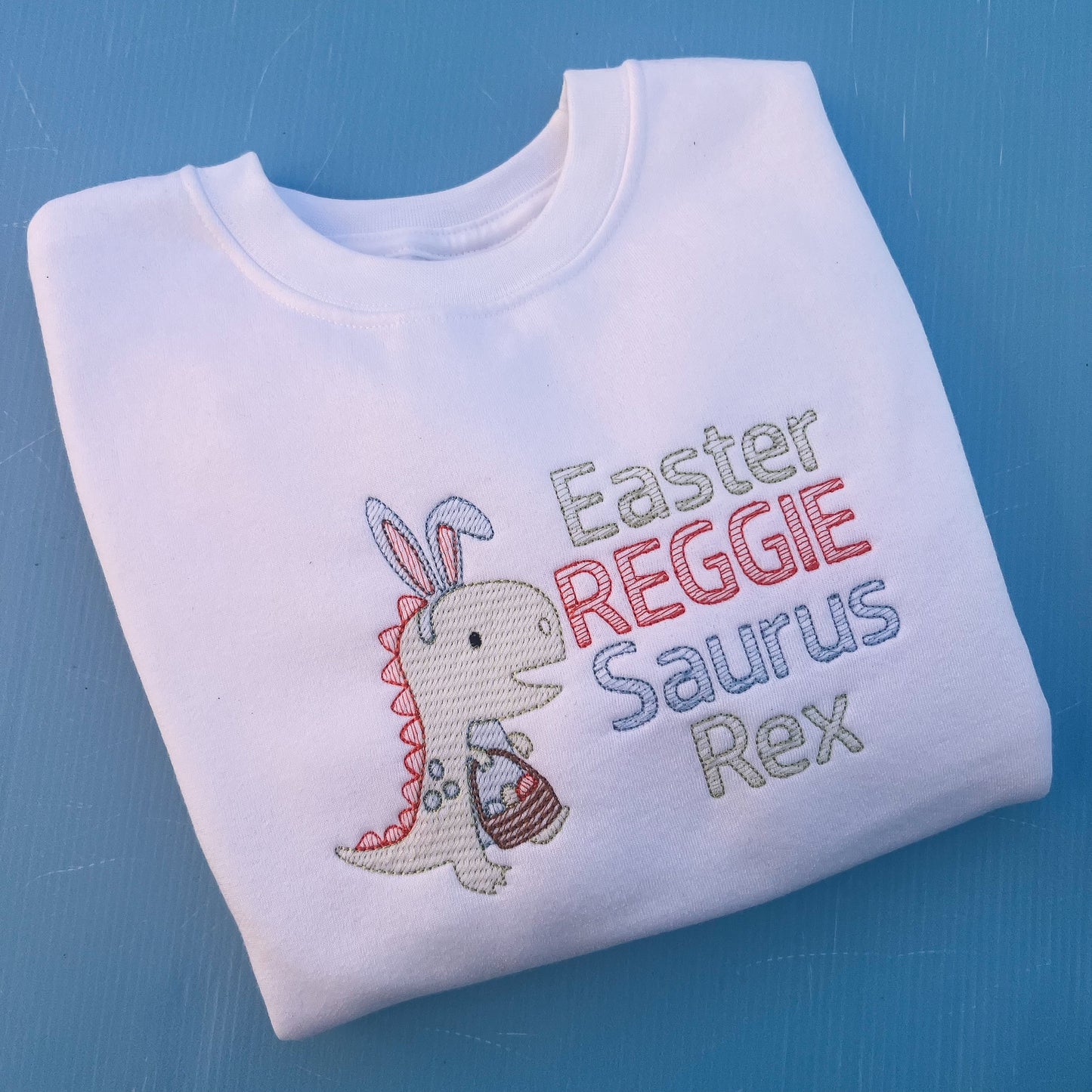 White sweatshirt with a dinosaur with bunny ears and a Easter egg basket with the words Easter Reggie Saurus Rex 