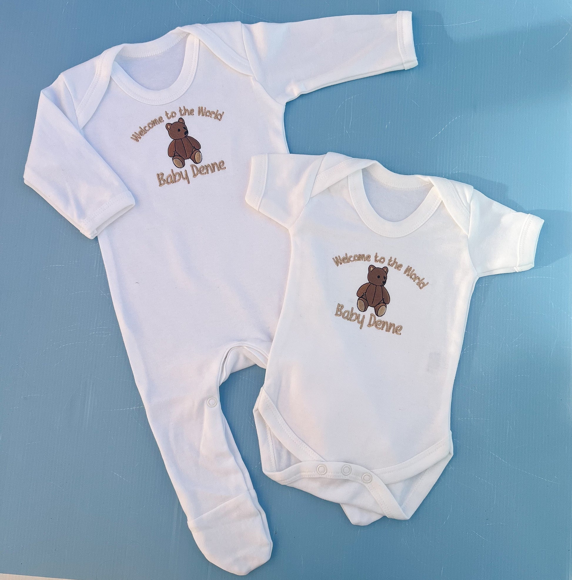Two baby onesies with embroidered text and a bear design on a blue background