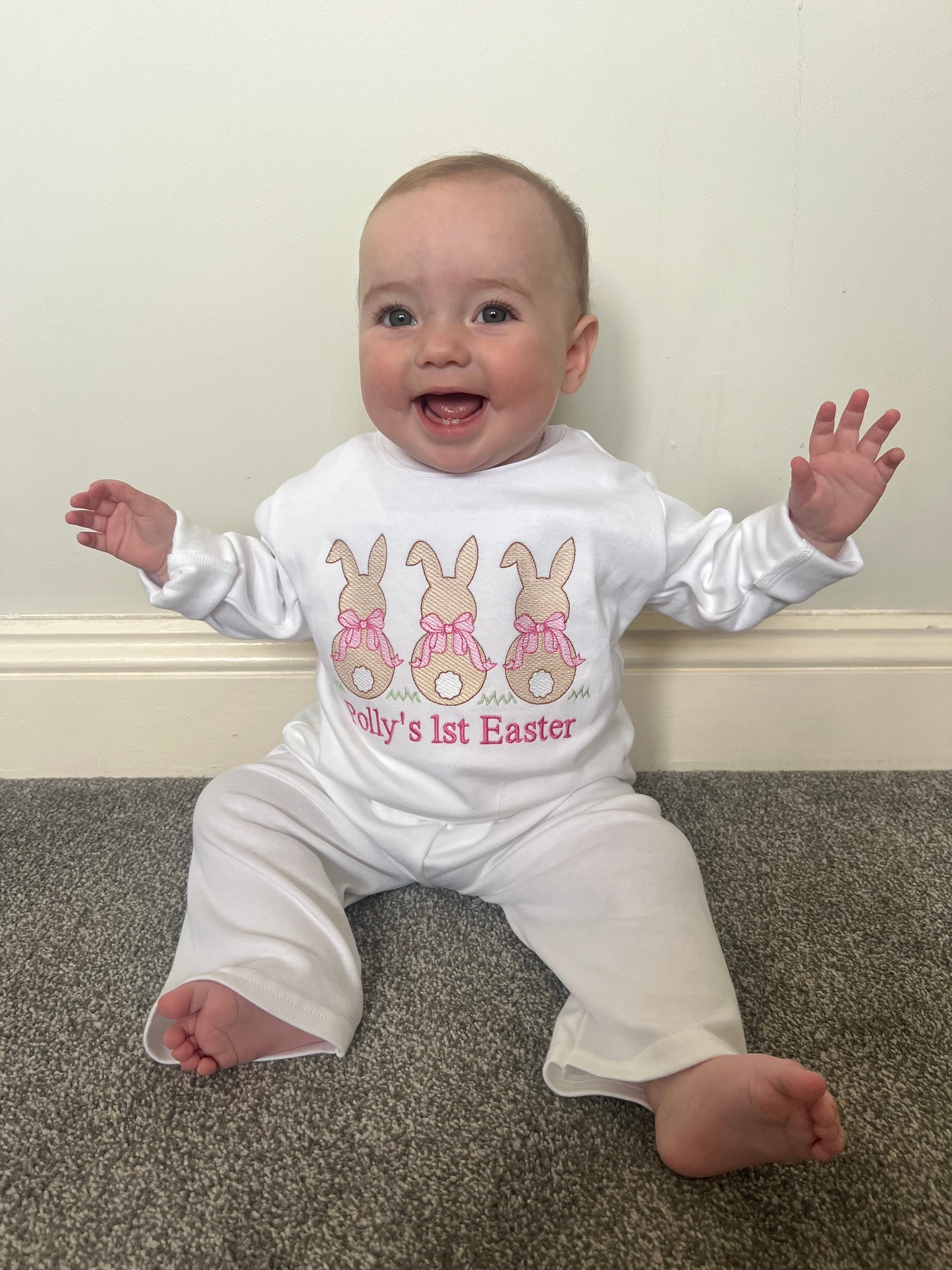 Baby wearing a 'My 1st Easter' onesie on a carpeted floor. 3 bunny’s with pink bows 