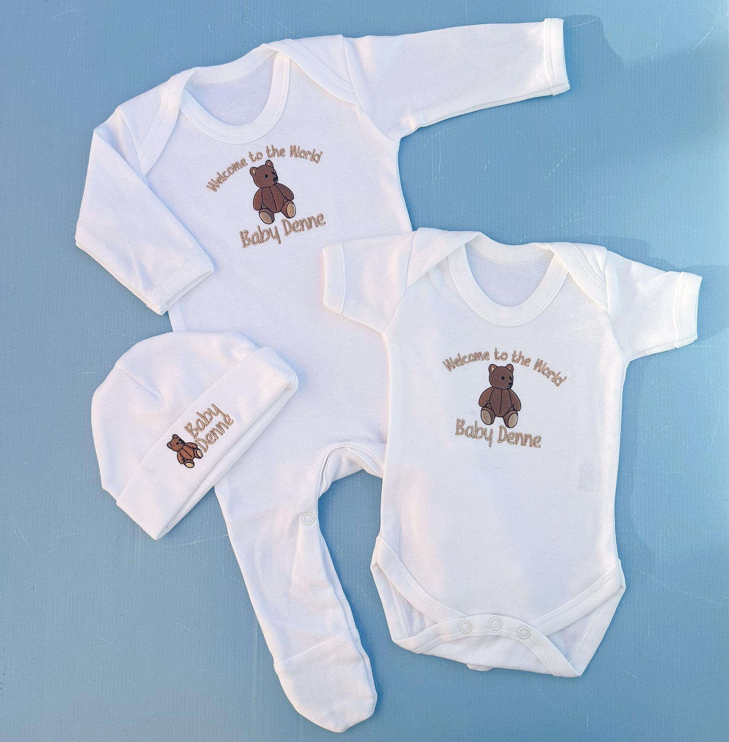 Baby clothing set with embroidered text and teddy bear design on a blue background