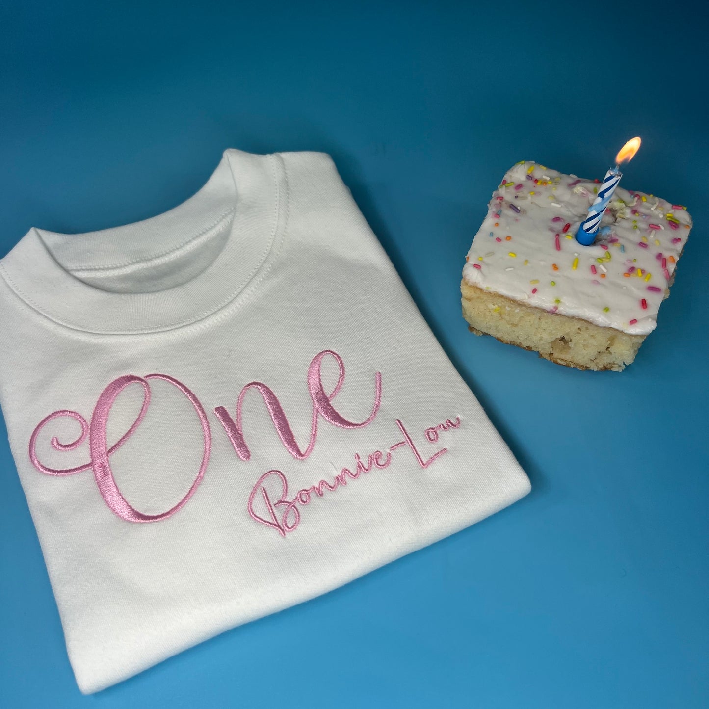 Personalised birthday t-shirt White shirt with 'One Bonnie Lou' text, next to a small cake with a candle on a blue background