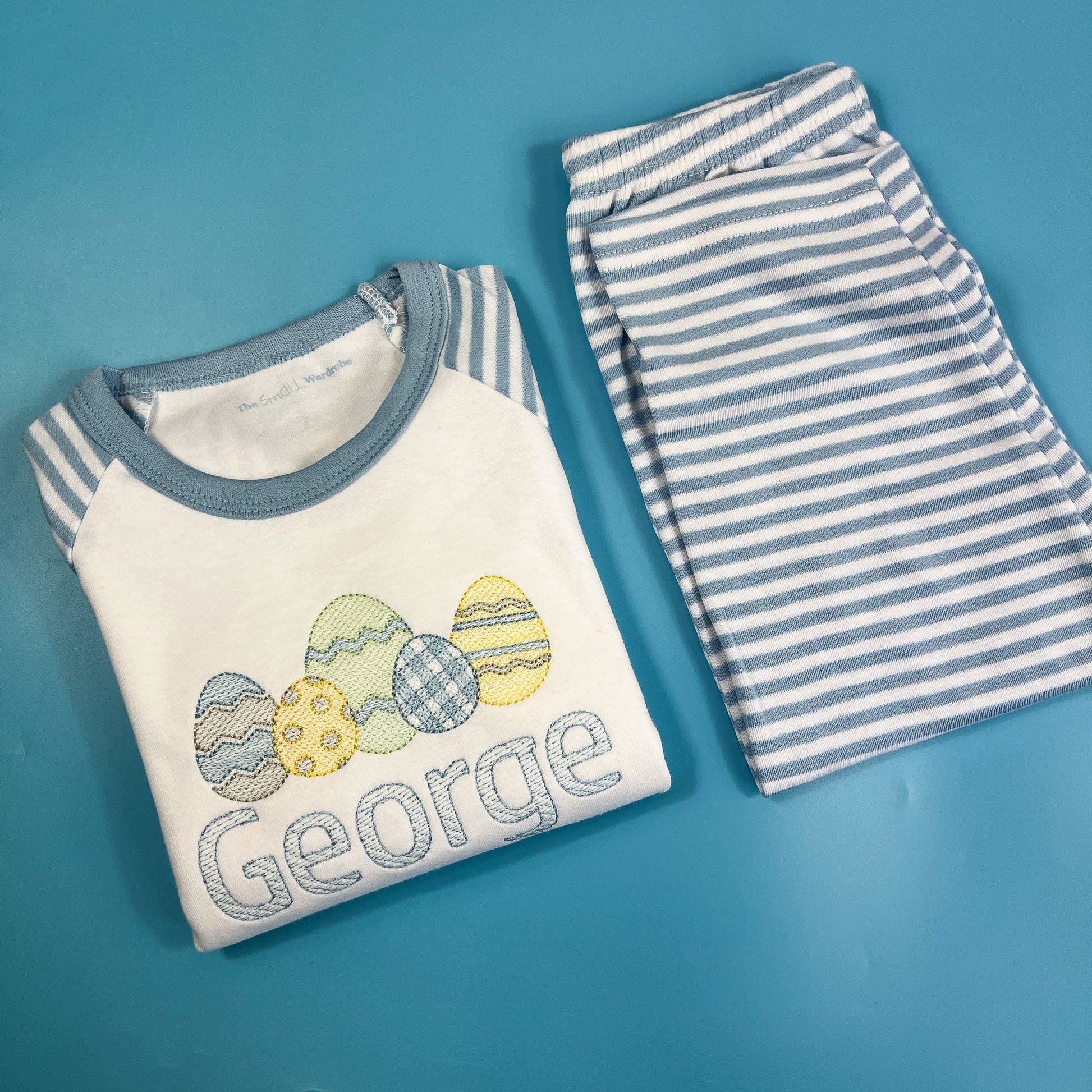 Children's shirt with embroidered eggs and 'George' text, paired with striped pants on a blue background