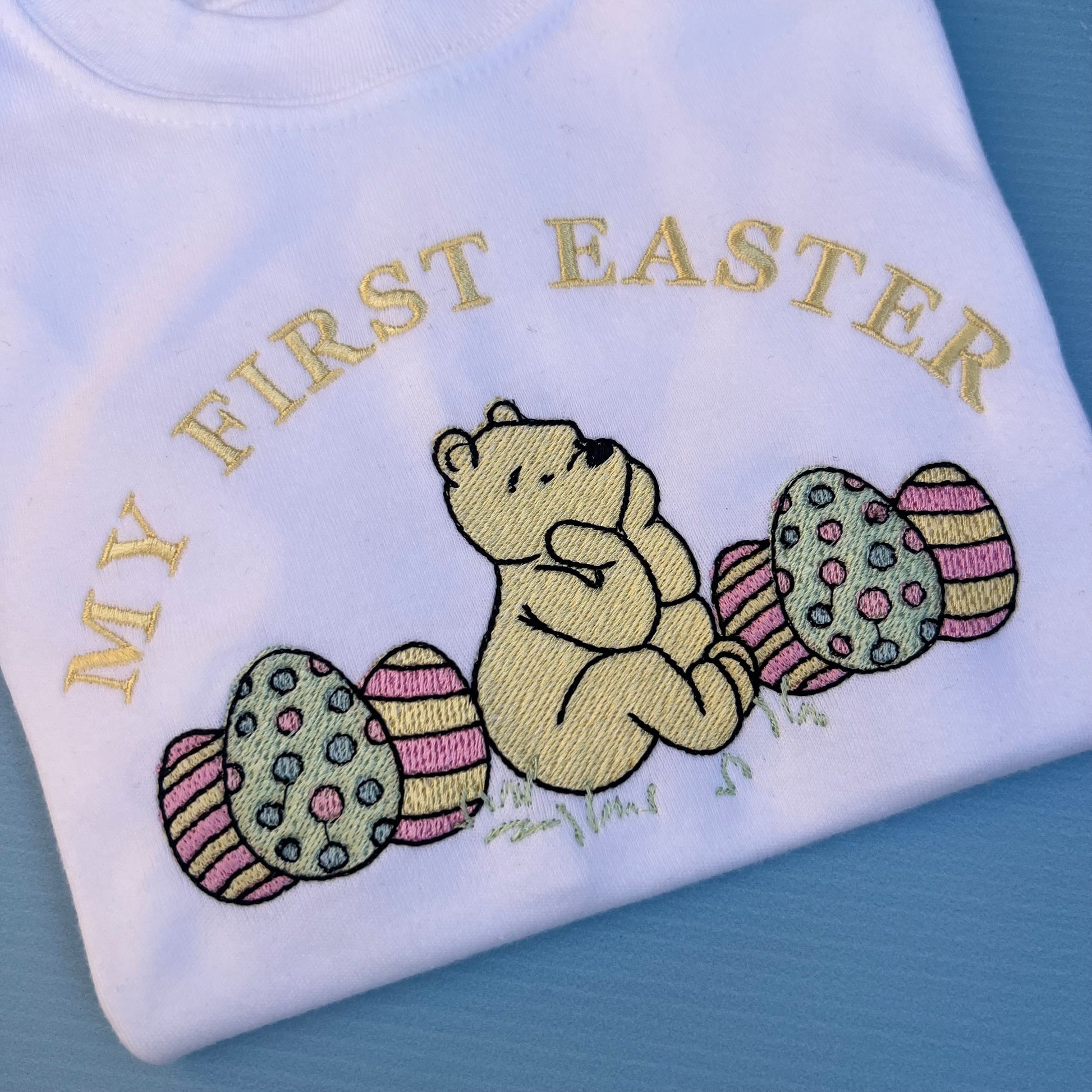 Embroidered design of a bear with Easter eggs on a white fabric with 'My First Easter' text.
