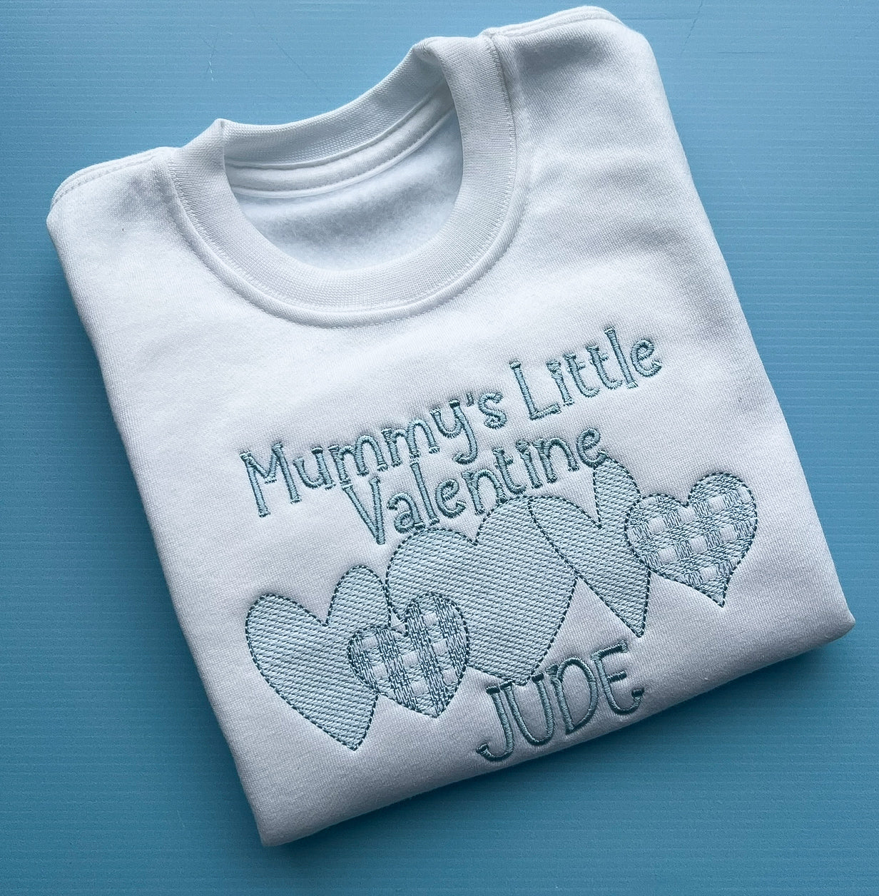 White sweatshirt with mummy’s little valentines embroidered in blue finished with a row of detailed hearts and the name Jude 