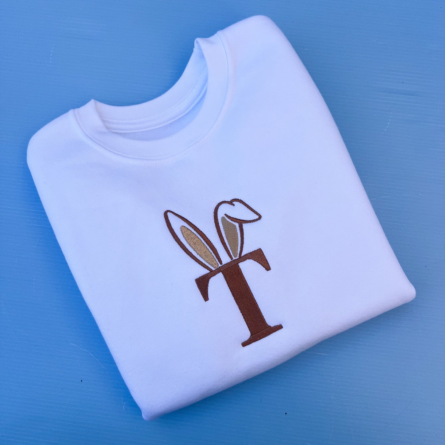 Folded white sweatshirt with a brown and beige initial with bunny ears on a blue background