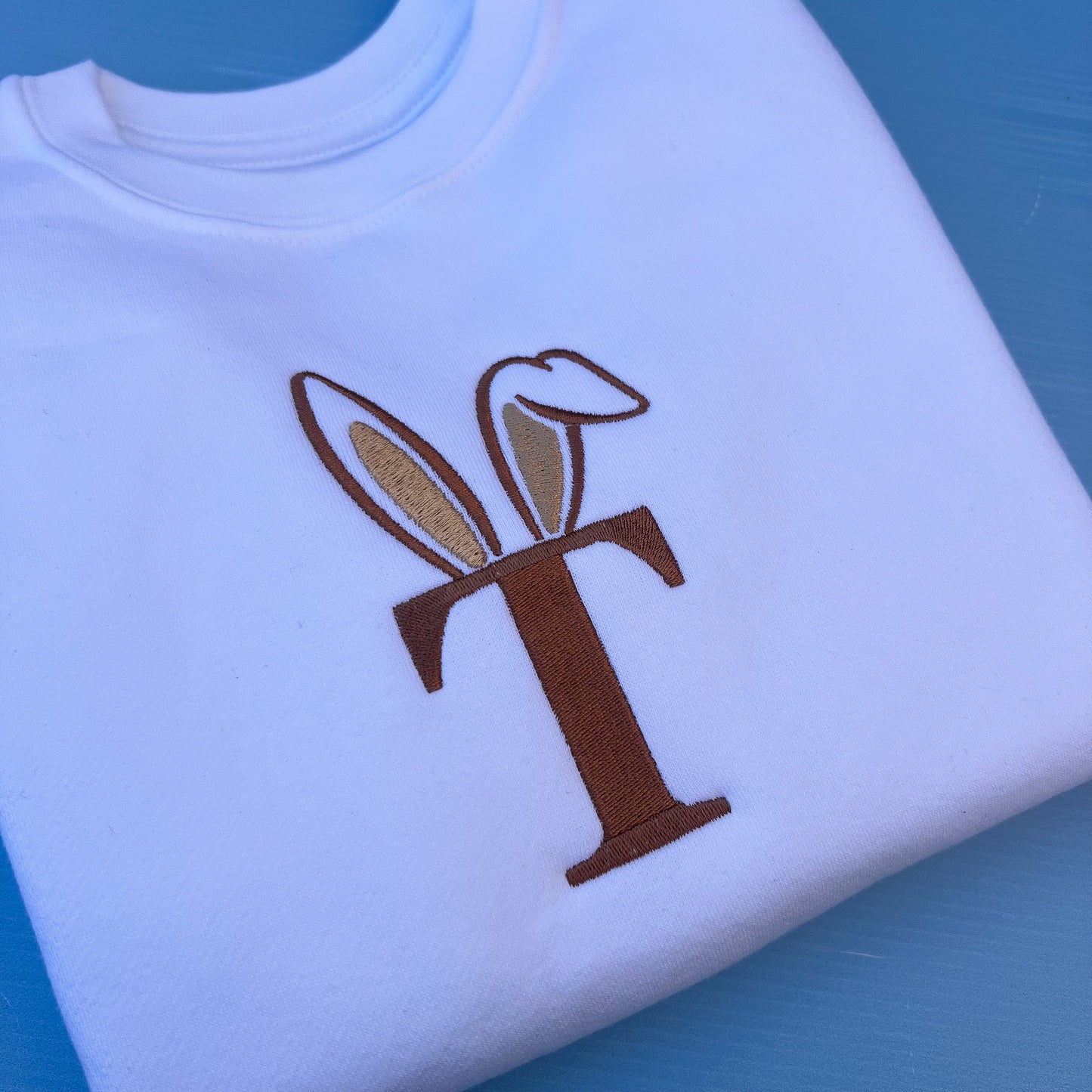 White sweatshirt with a brown initial with bunny ears on a blue background
