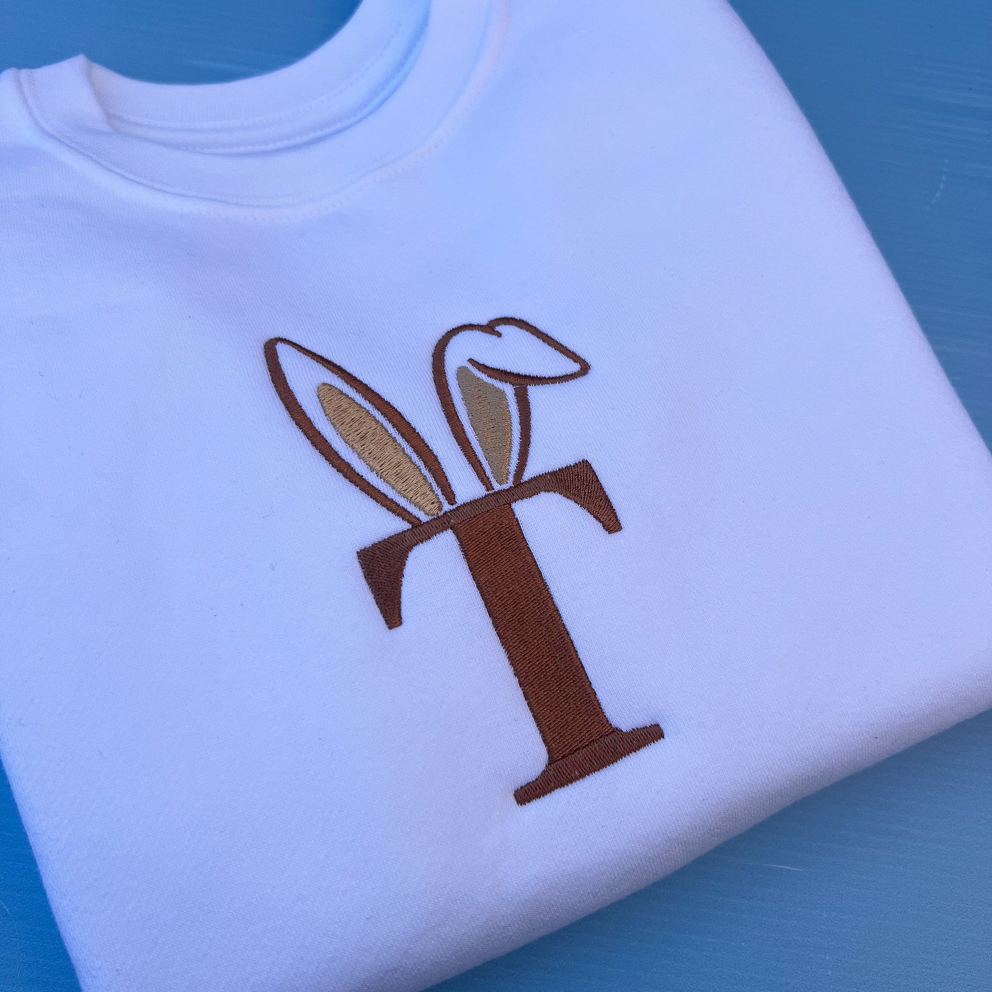 White sweatshirt with a brown initial with bunny ears on a blue background