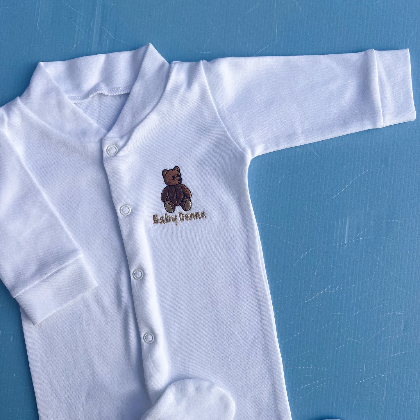 White baby onesie with a brown bear design and 'Baby Denne' text on a blue background