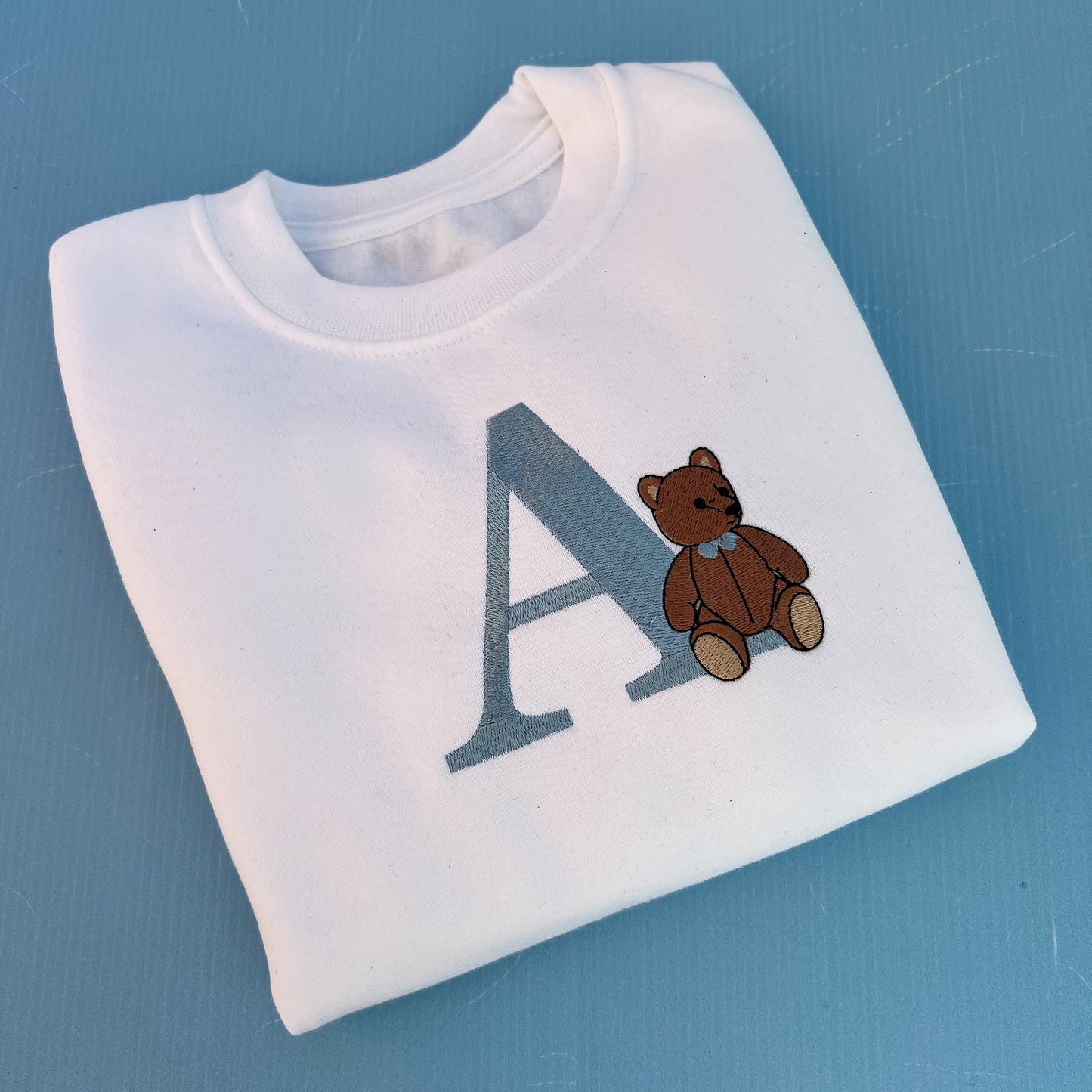 White sweatshirt with a teddy bear and letter 'A' on a blue background