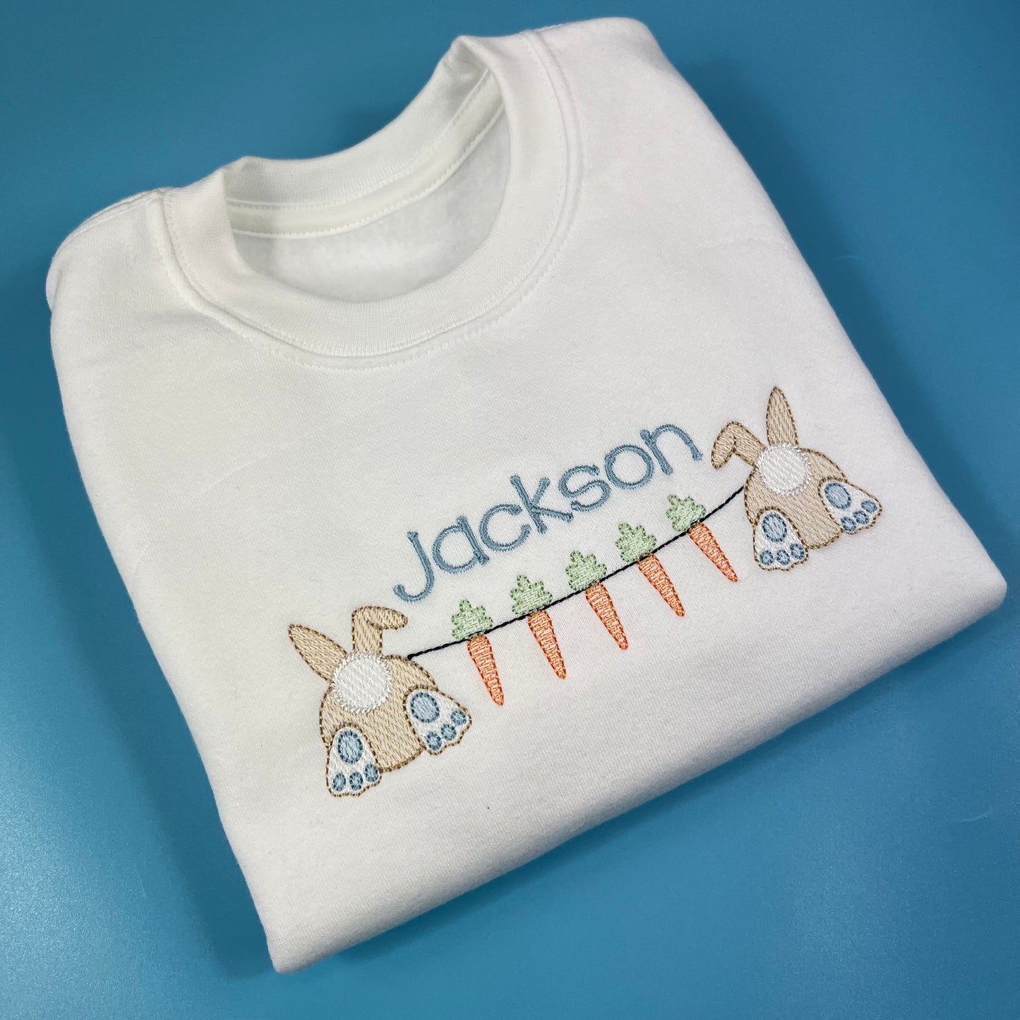 Folded white sweatshirt with embroidered name and bunny design on a blue background