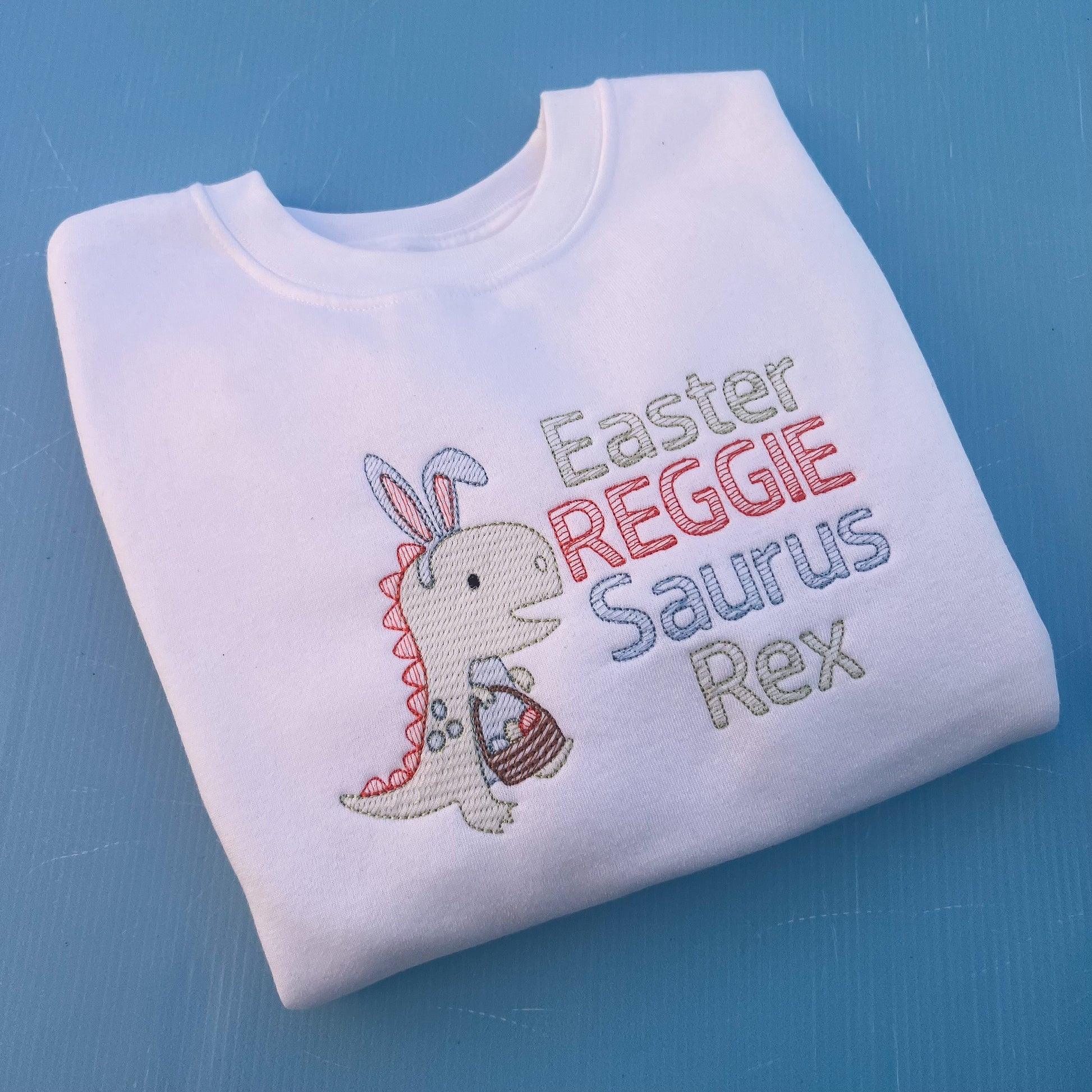 White sweatshirt with a dinosaur with bunny ears and a Easter egg basket with the words Easter Reggie Saurus Rex 