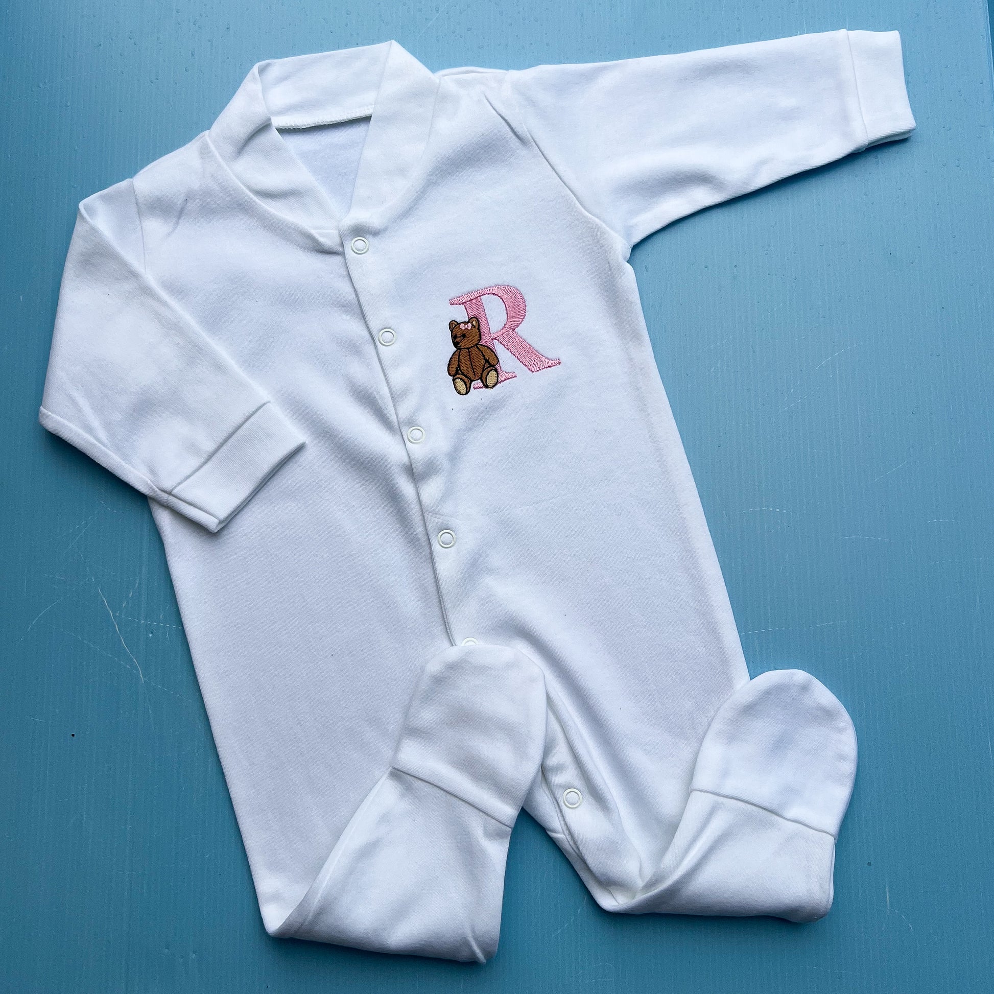 White baby onesie with a traditional teddy bear and letter 'R' on a blue background