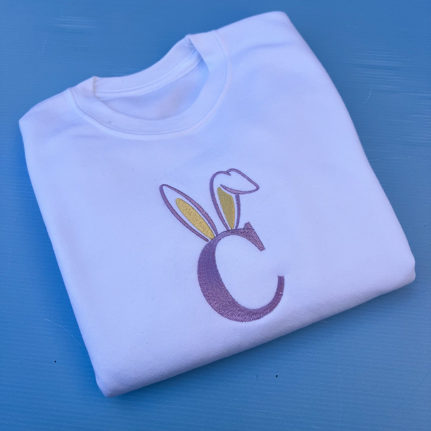 White t-shirt with a purple letter 'C' and bunny ears design on a blue background