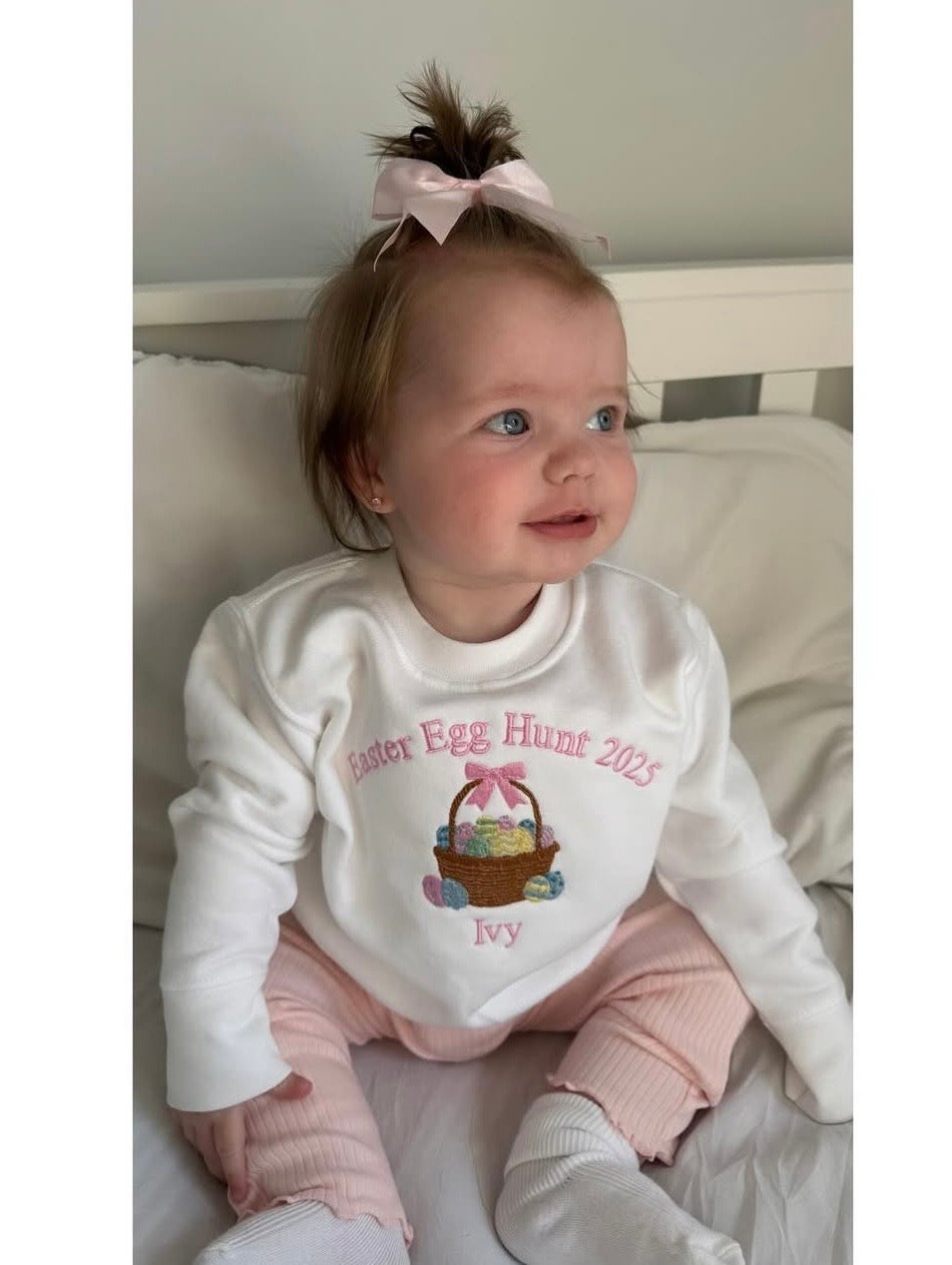 Baby wearing a white sweatshirt with 'Easter Egg Hunt 2023' and 'Ivy' text, sitting on a bed.