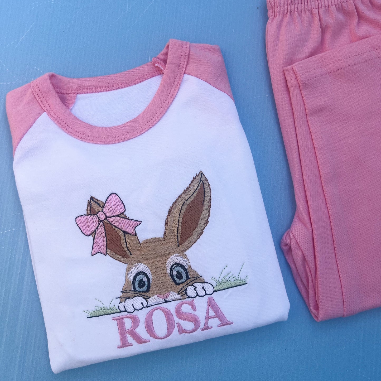 Pink pyjamas with a bun peaking over some grass with a bow on her ear. Finished off with the name Rosa. 