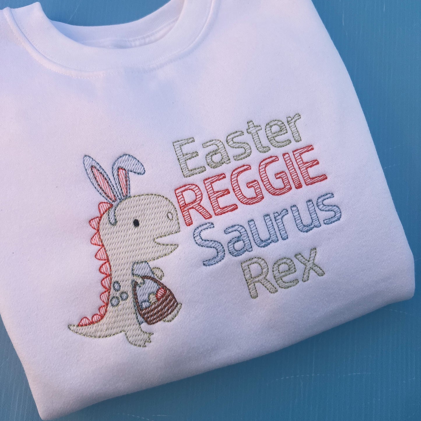 White sweatshirt with a dinosaur with bunny ears and a Easter egg basket with the words Easter Reggie Saurus Rex 