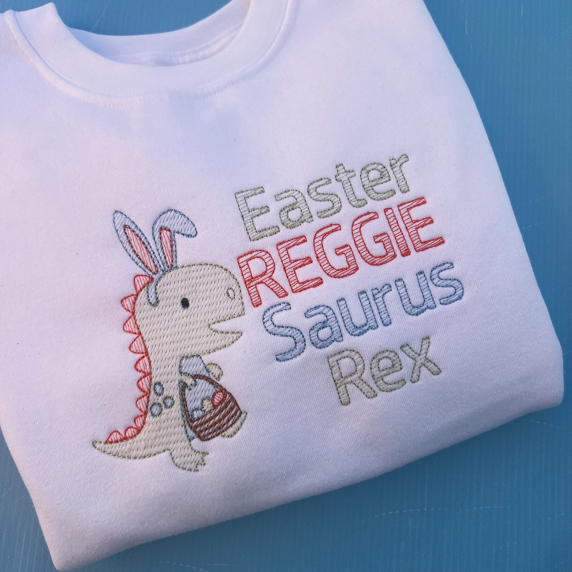 White sweatshirt with a dinosaur with bunny ears and a Easter egg basket with the words Easter Reggie Saurus Rex 