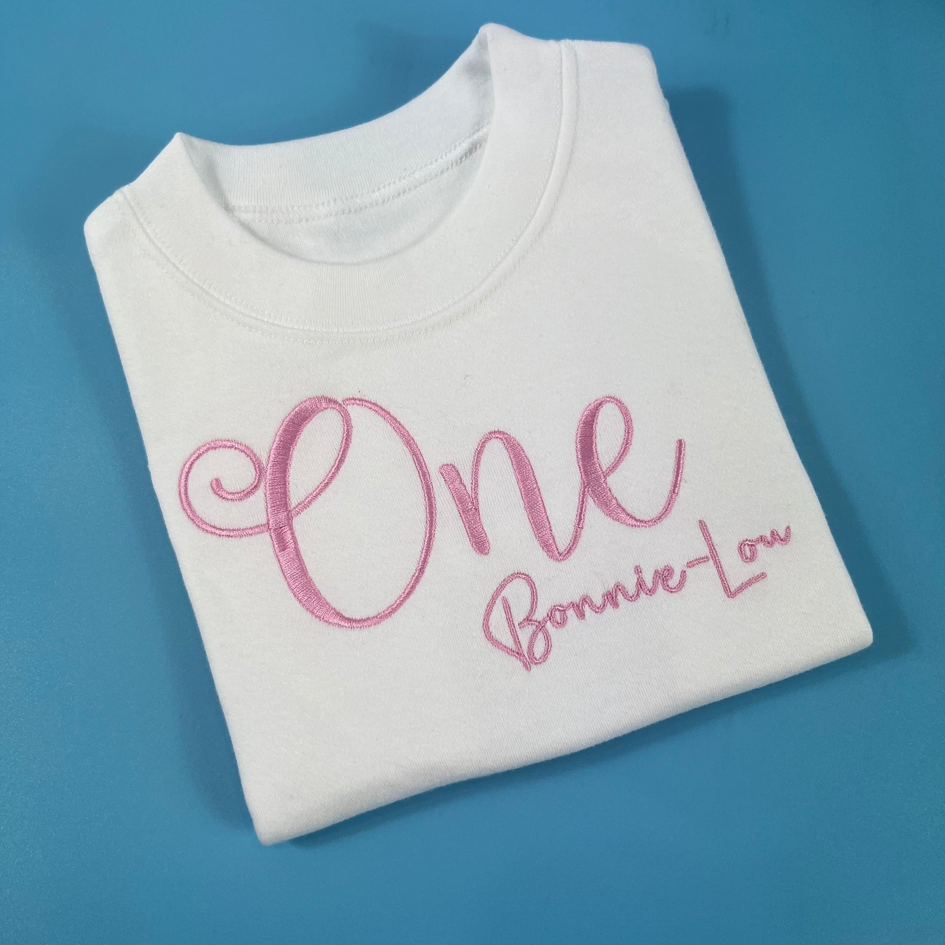 Folded white sweatshirt with pink text on a blue background. Personalised birthday t-shirt 