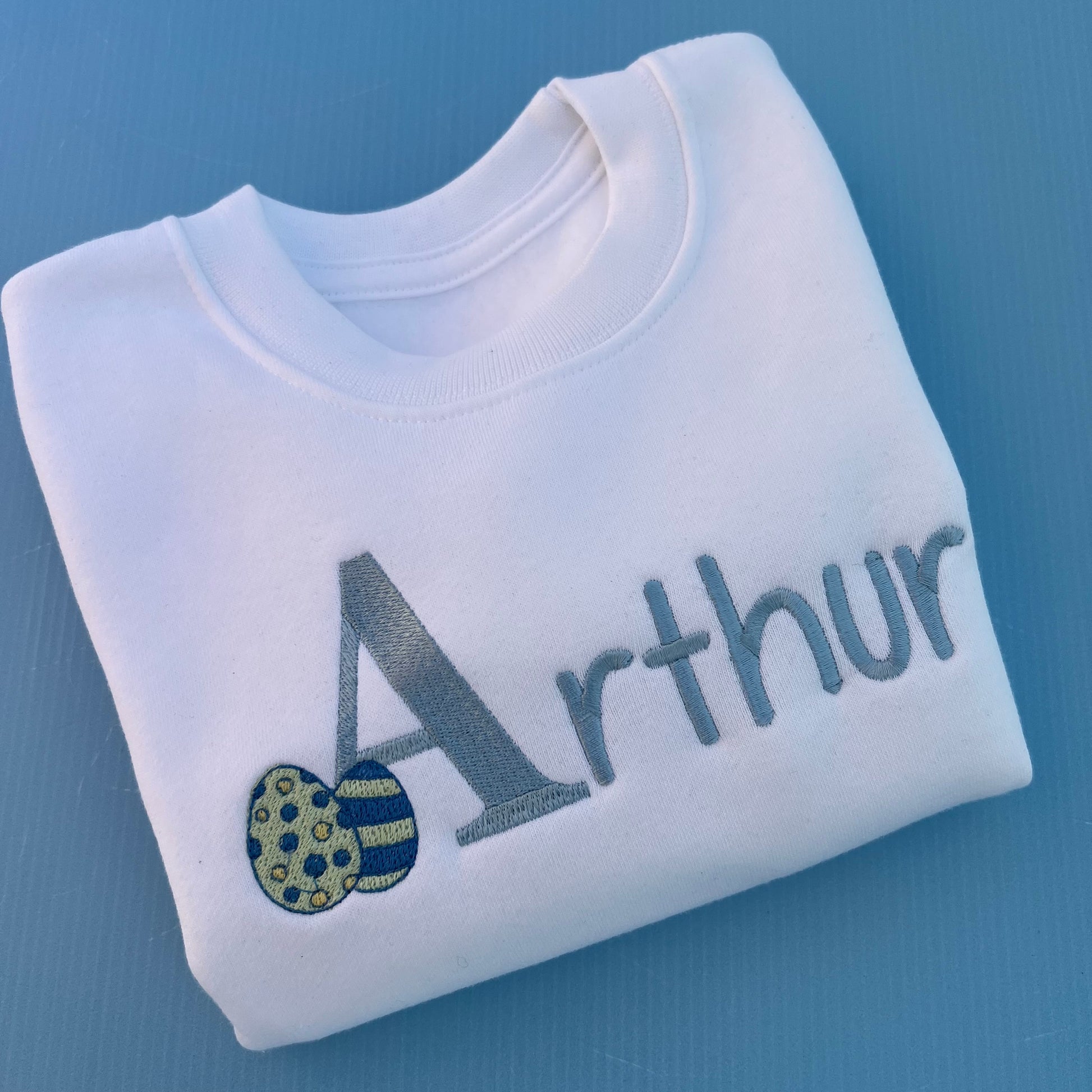 Folded white sweatshirt with 'Arthur' embroidered on it with 2 little Easter eggs against a blue background