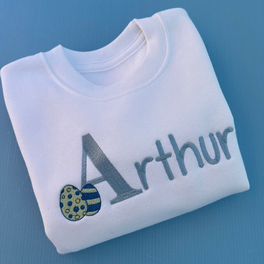 Folded white sweatshirt with 'Arthur' embroidered on it with 2 little Easter eggs against a blue background