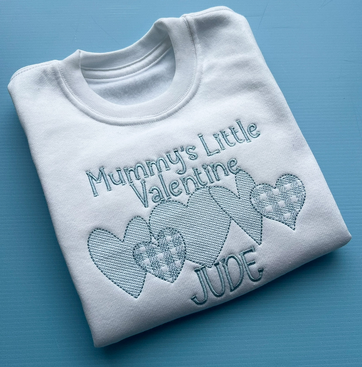 White sweatshirt with mummy’s little valentines embroidered in blue finished with a row of detailed hearts and the name Jude 